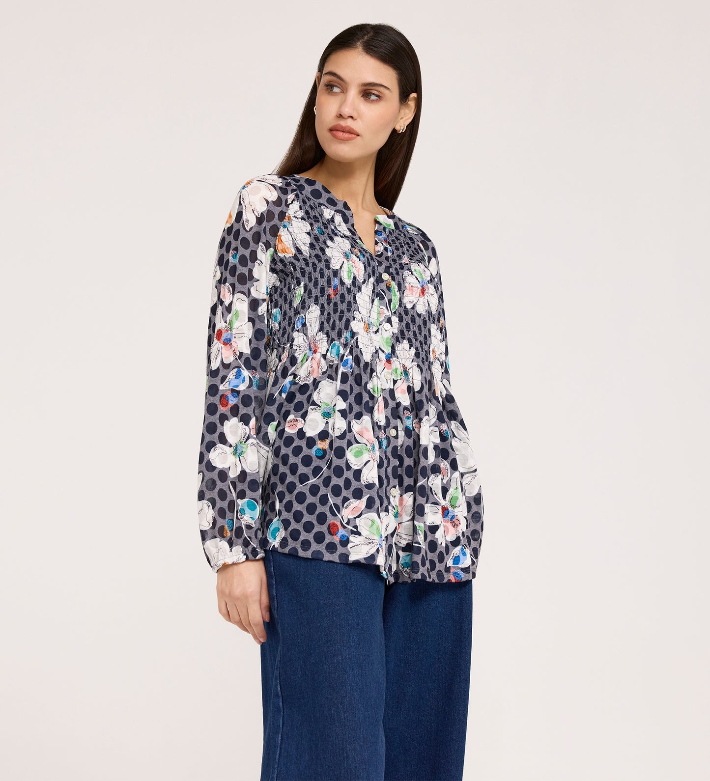 A woman in a Martha Navy Flower Jersey Blouse with textured shirring, floral and polka dot patterns, long sleeves, and blue wide-leg jeans stands against a white background, looking slightly left with a neutral expression.