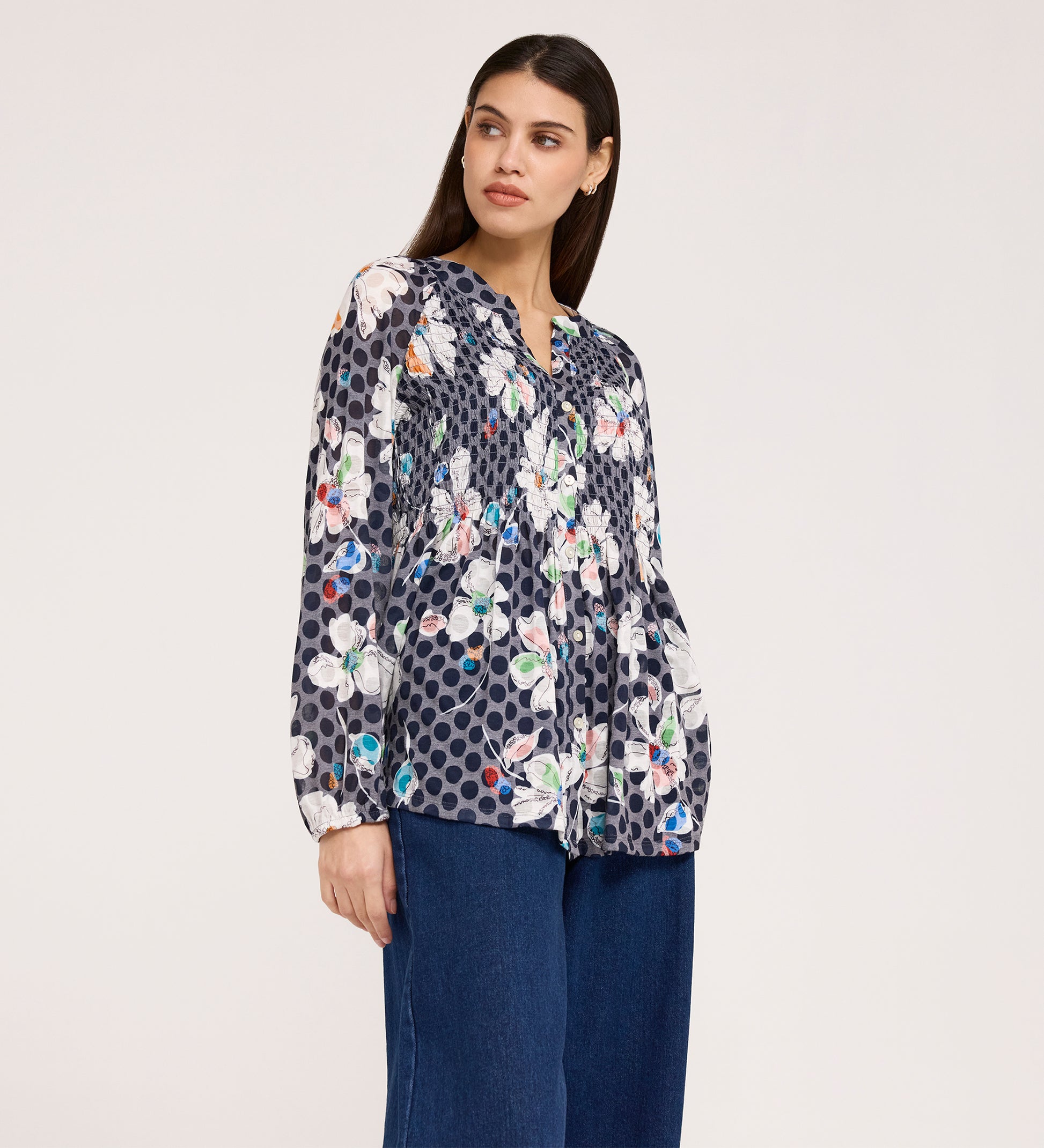 A woman in a Martha Navy Flower Jersey Blouse with textured shirring, floral and polka dot patterns, long sleeves, and blue wide-leg jeans stands against a white background, looking slightly left with a neutral expression.