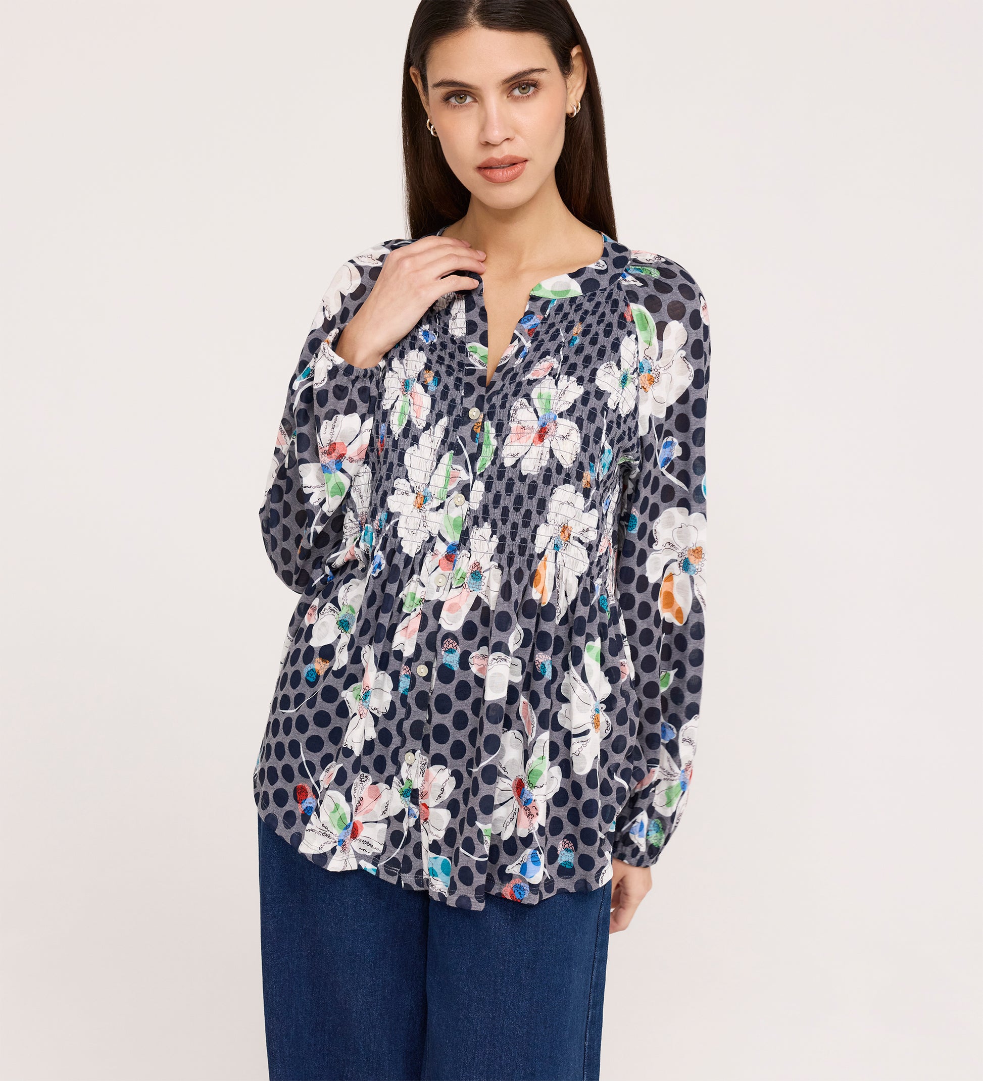 A woman with long dark hair wears the Martha Navy Flower Jersey Blouse, a navy long-sleeve top with white floral and polka dot patterns and shirring, paired with blue wide-leg pants, against a plain light background.