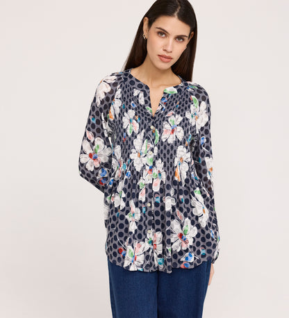 A woman with long dark hair wears the Martha Navy Flower Jersey Blouse, which has a navy base with white, pink, and orange floral prints plus dot patterns, a subtle flare hem, and textured shirring. She poses against a plain light background.