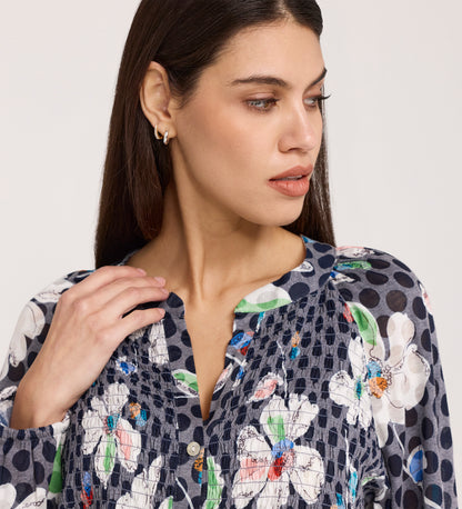 A woman with straight dark hair looks to the side, wearing small hoop earrings and a Martha Navy Flower Jersey Blouse with white floral and polka dot patterns, a subtle flare hem, and one hand near her collarbone.