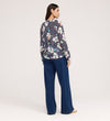 Woman standing and facing away, wearing a long-sleeve Martha Navy Flower Jersey Blouse with textured shirring and wide-leg blue jeans against a plain, light-colored background.