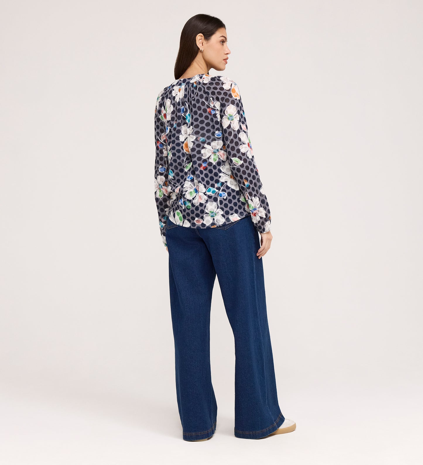 Woman standing and facing away, wearing a long-sleeve Martha Navy Flower Jersey Blouse with textured shirring and wide-leg blue jeans against a plain, light-colored background.