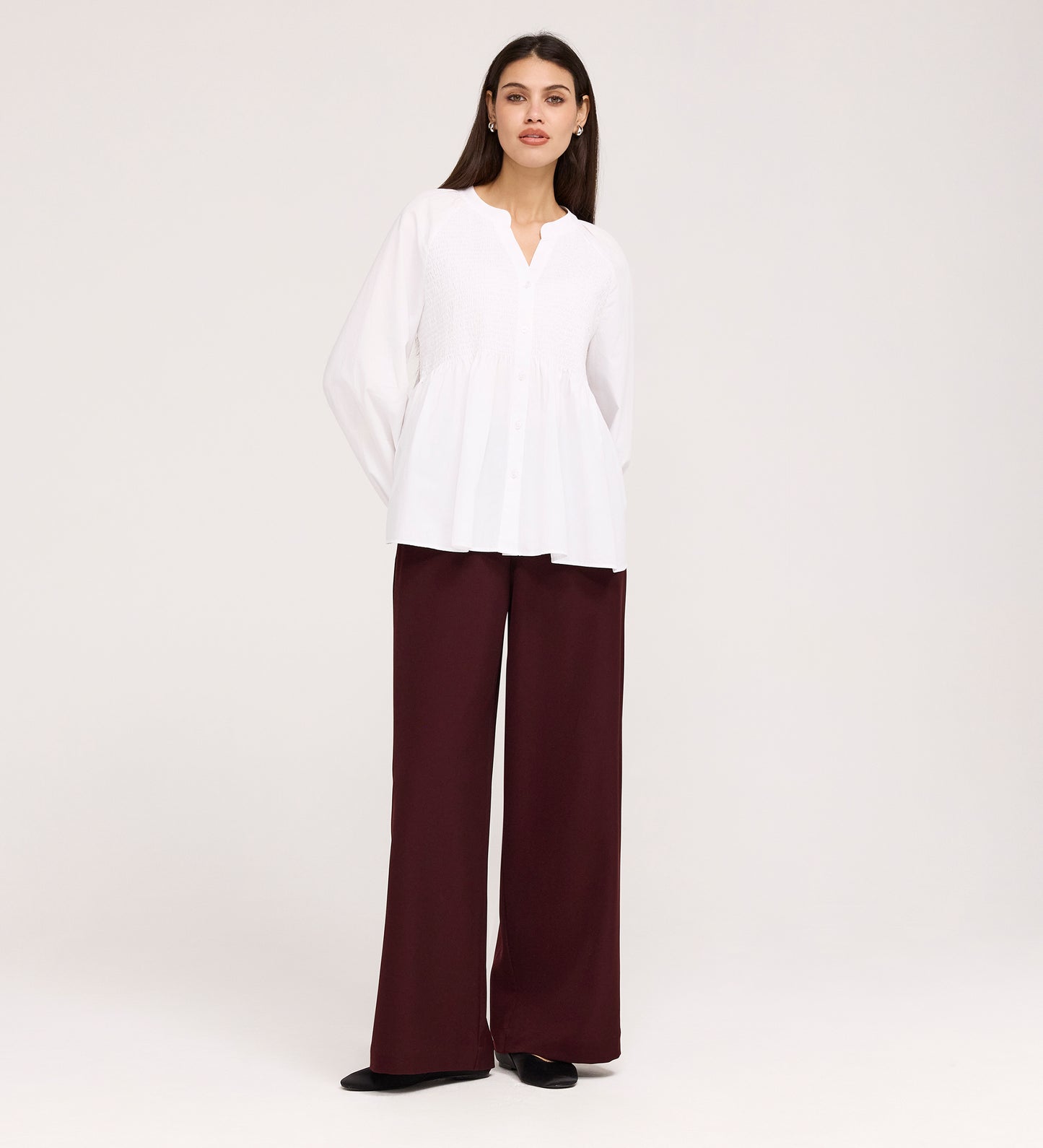 A woman stands against a plain background wearing the Martha White Cotton Blouse with a v-neck and long sleeves, paired with high-waisted, wide-leg maroon pants and black shoes.