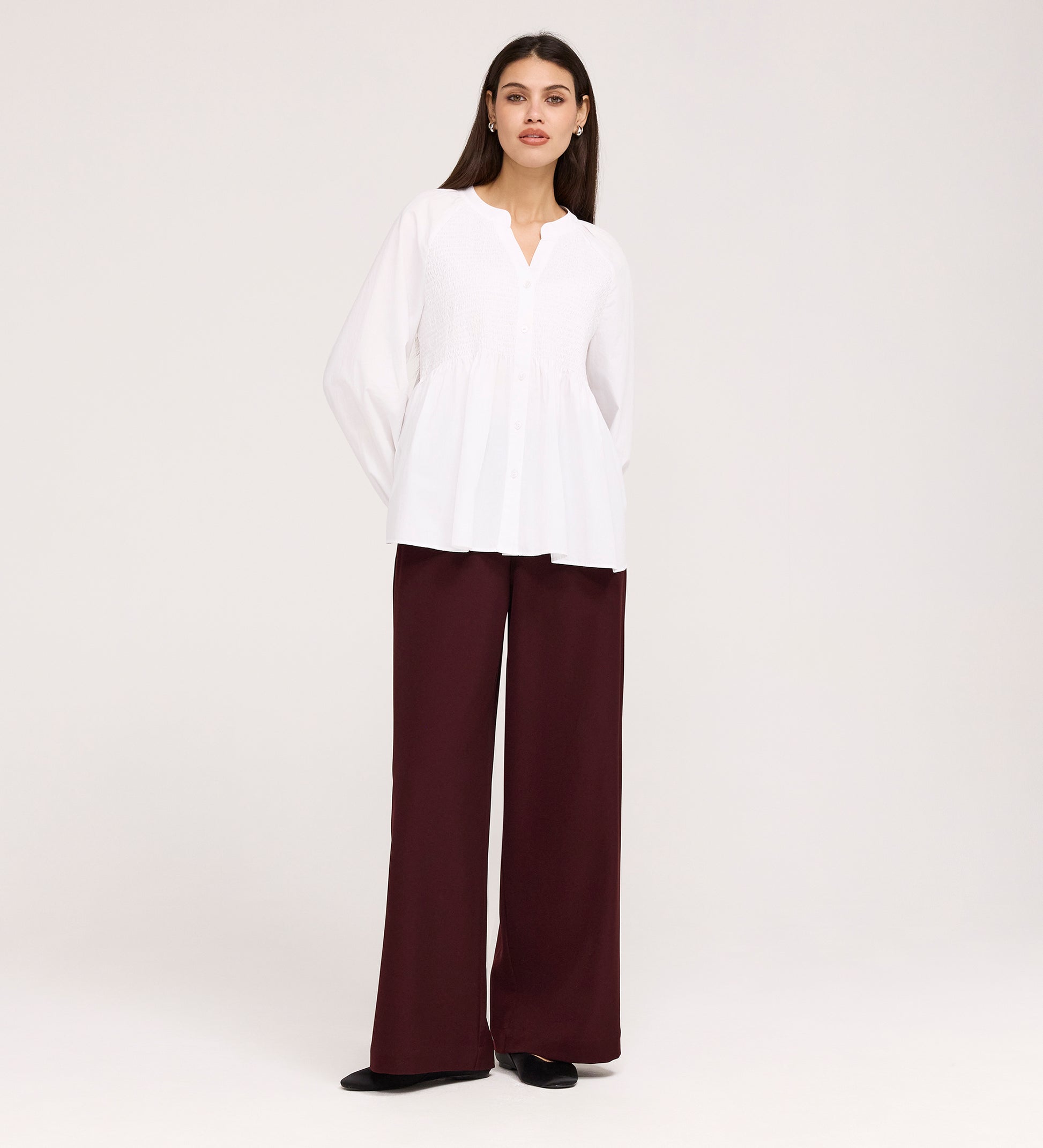 A woman stands against a plain background wearing the Martha White Cotton Blouse with a v-neck and long sleeves, paired with high-waisted, wide-leg maroon pants and black shoes.