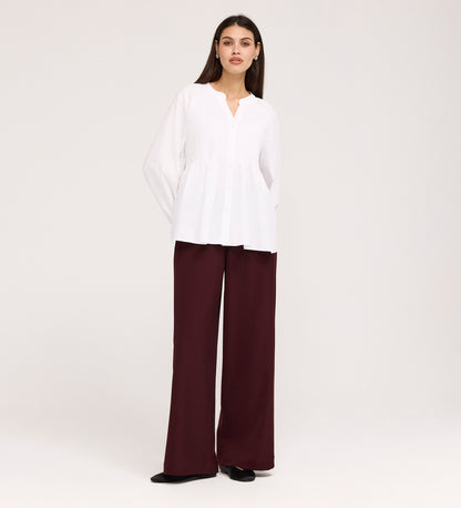 A woman stands against a plain background wearing the Martha White Cotton Blouse with a v-neck and long sleeves, paired with high-waisted, wide-leg maroon pants and black shoes.