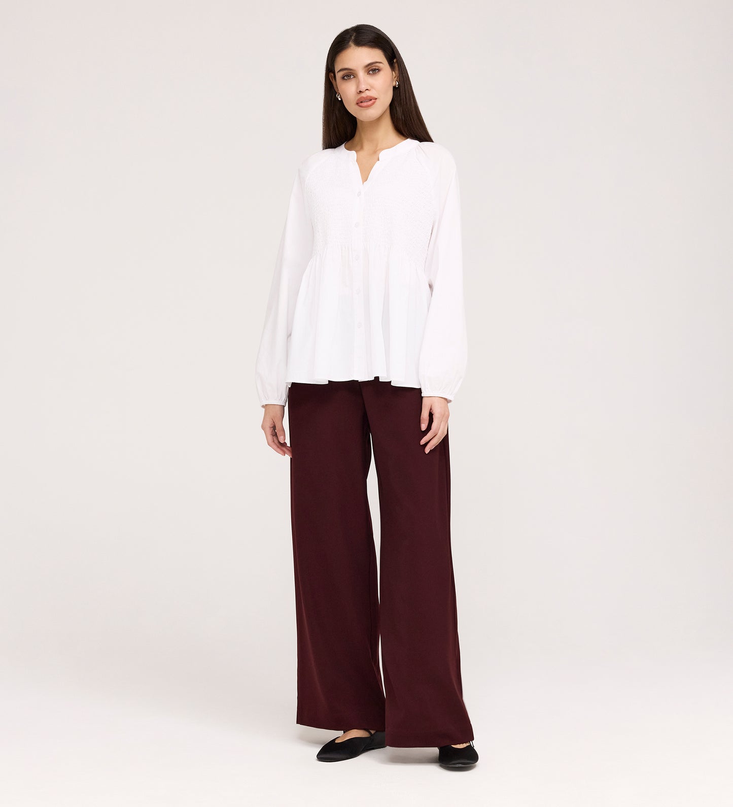 A woman with straight dark hair faces forward, wearing a Martha White Cotton Blouse with buttons and loose maroon wide-leg pants. She pairs the outfit with black flats and poses against a plain light background.