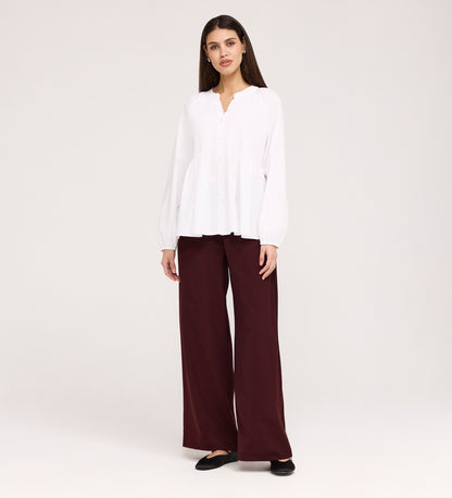 A woman with straight dark hair faces forward, wearing a Martha White Cotton Blouse with buttons and loose maroon wide-leg pants. She pairs the outfit with black flats and poses against a plain light background.