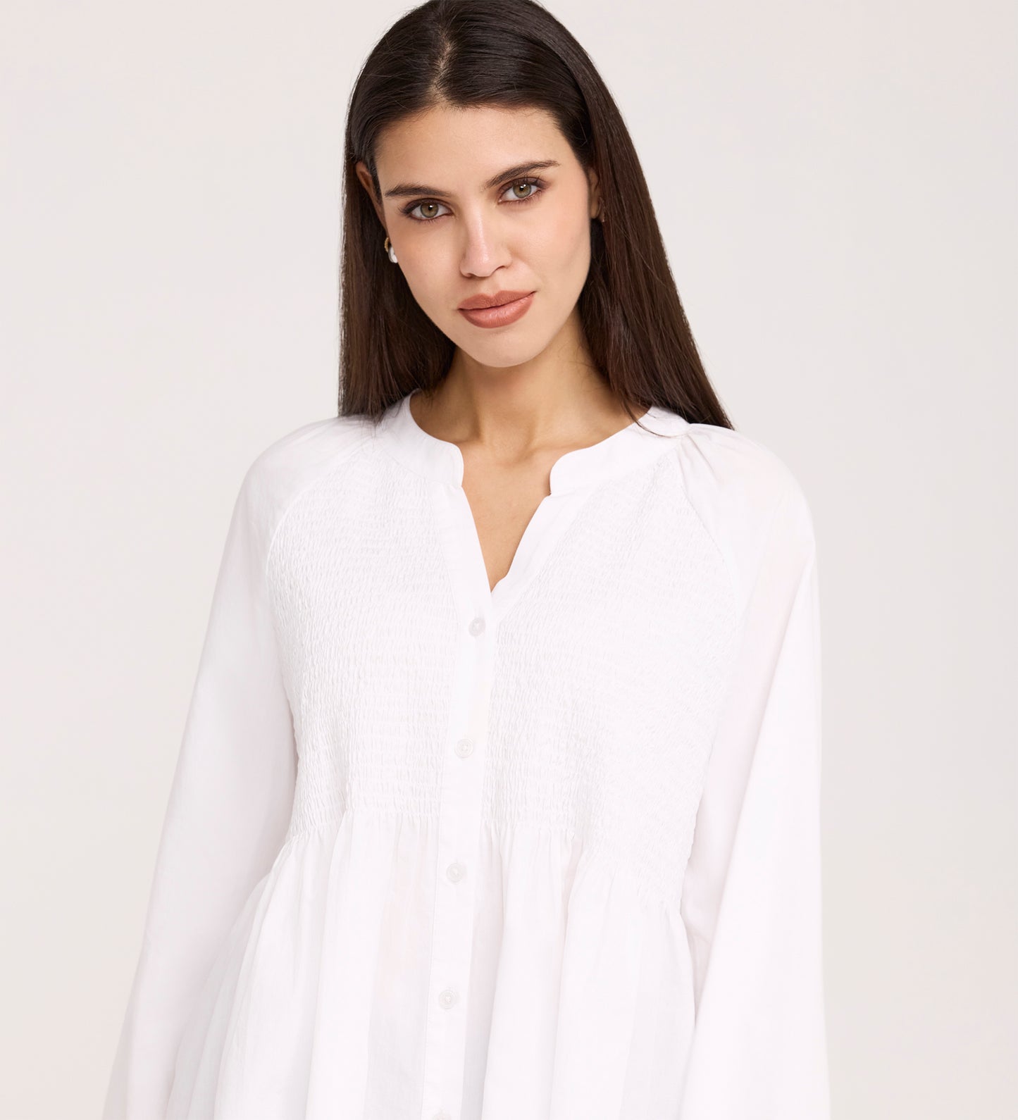 A woman with long dark hair wears a loose-fitting Martha White Cotton Blouse with a gathered bodice and buttons. The peasant-style top stands out against a light background as she gazes at the camera.