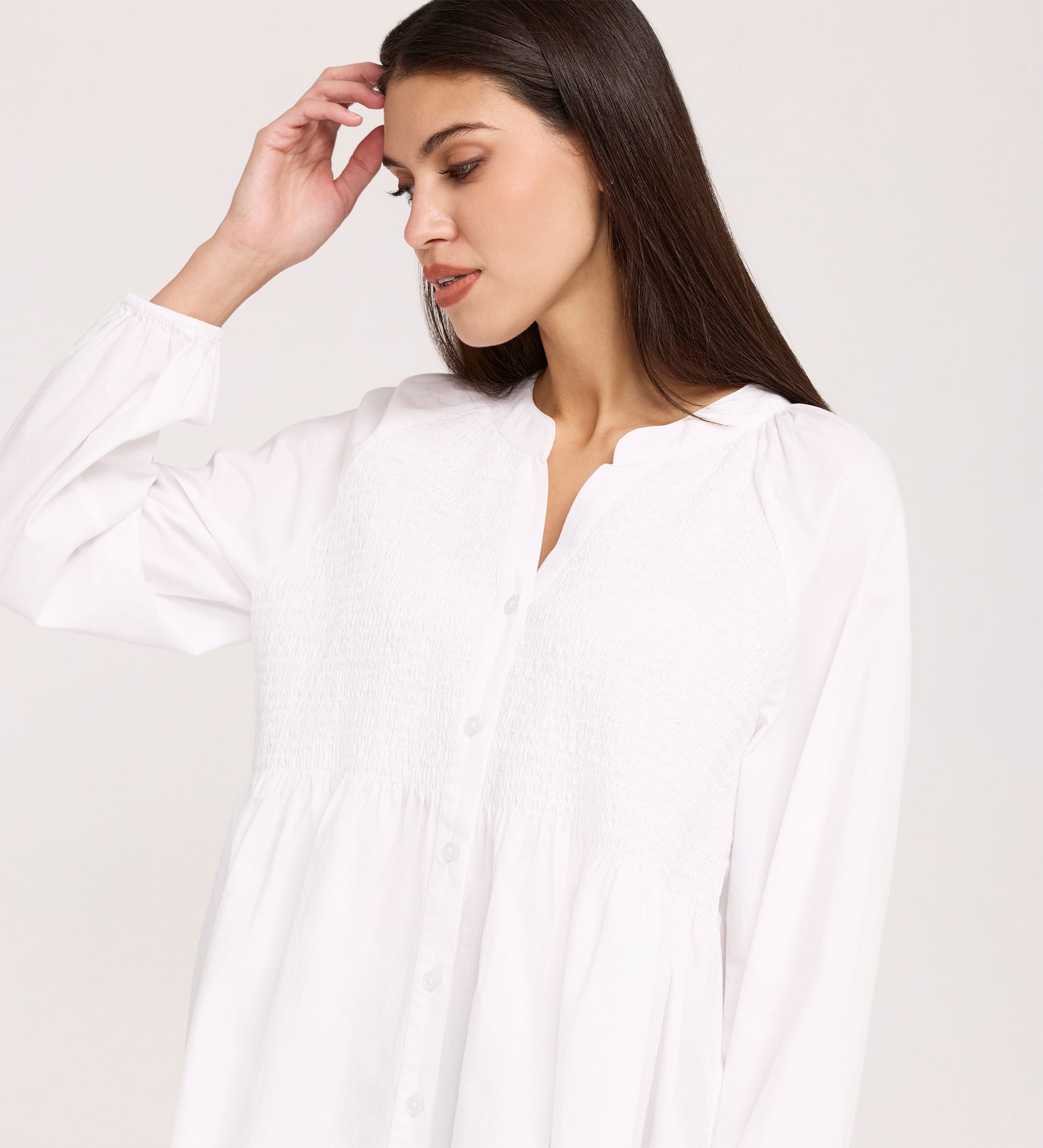 A woman with long dark hair wears a loose-fitting Martha White Cotton Blouse with a textured smocked panel on the upper bodice. She looks down and gently touches her hair against a plain background.