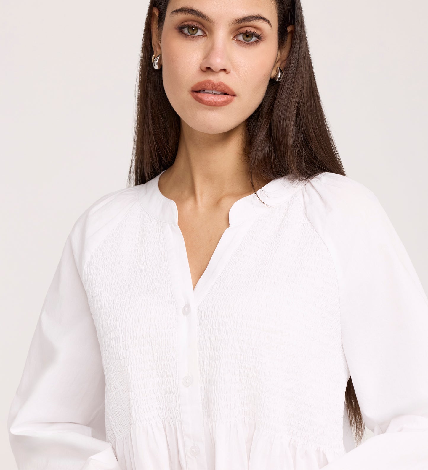 A woman with long brown hair, wearing silver hoop earrings and the Martha White Cotton Blouse with a smocked panel bodice, looks slightly to the side with a neutral expression.