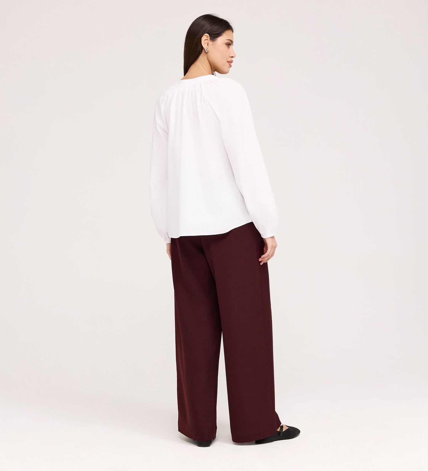 A woman stands facing away, wearing the Martha White Cotton Blouse with a smocked panel and wide-leg maroon pants, paired with black flats against a plain light background.