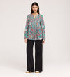 A woman stands facing forward in the Martha Green Floral Jersey Blouse with textured shirring, loose black flare-hem pants, and beige shoes, set against a plain light background.