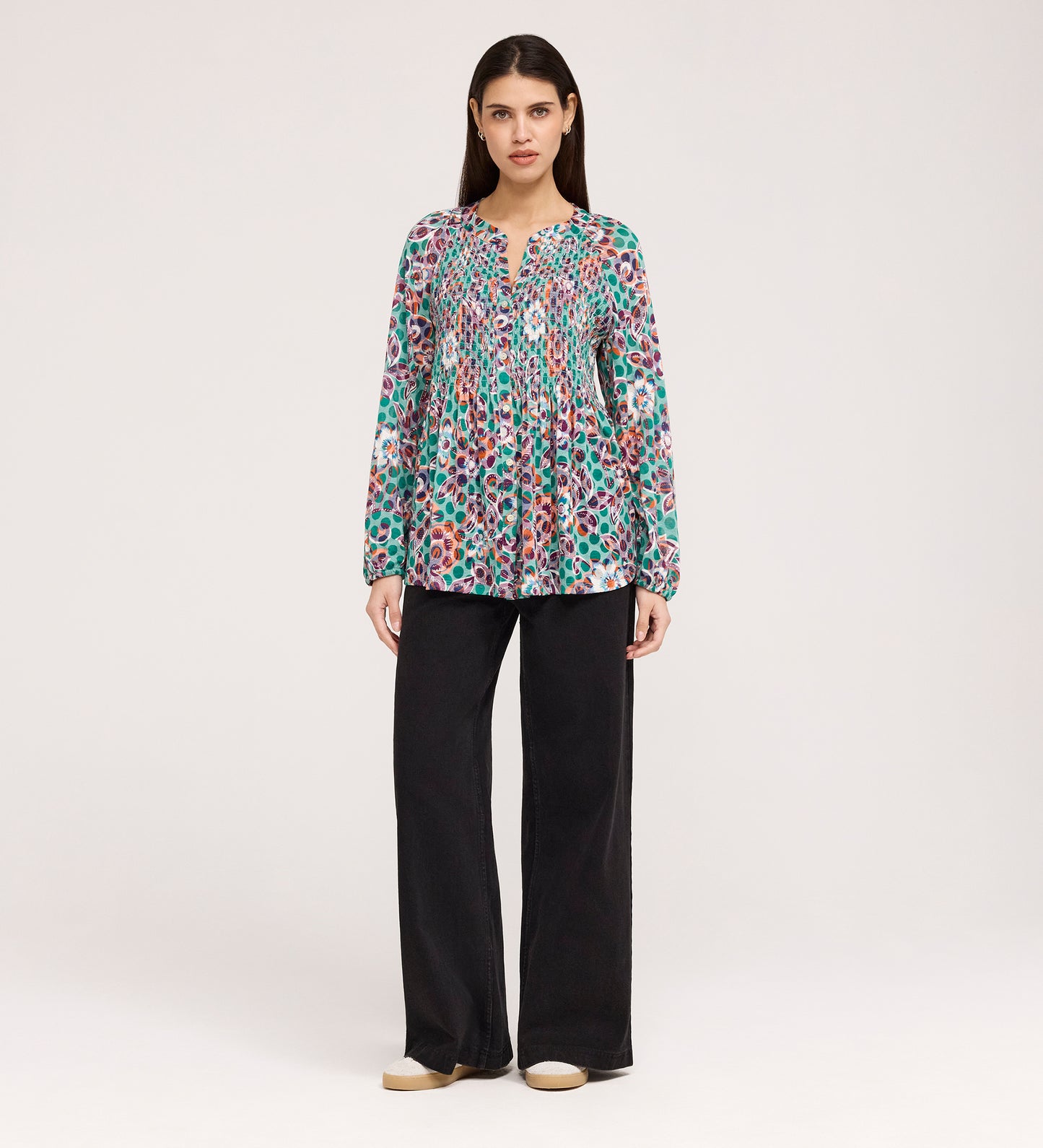 A woman stands facing forward in the Martha Green Floral Jersey Blouse with textured shirring, loose black flare-hem pants, and beige shoes, set against a plain light background.