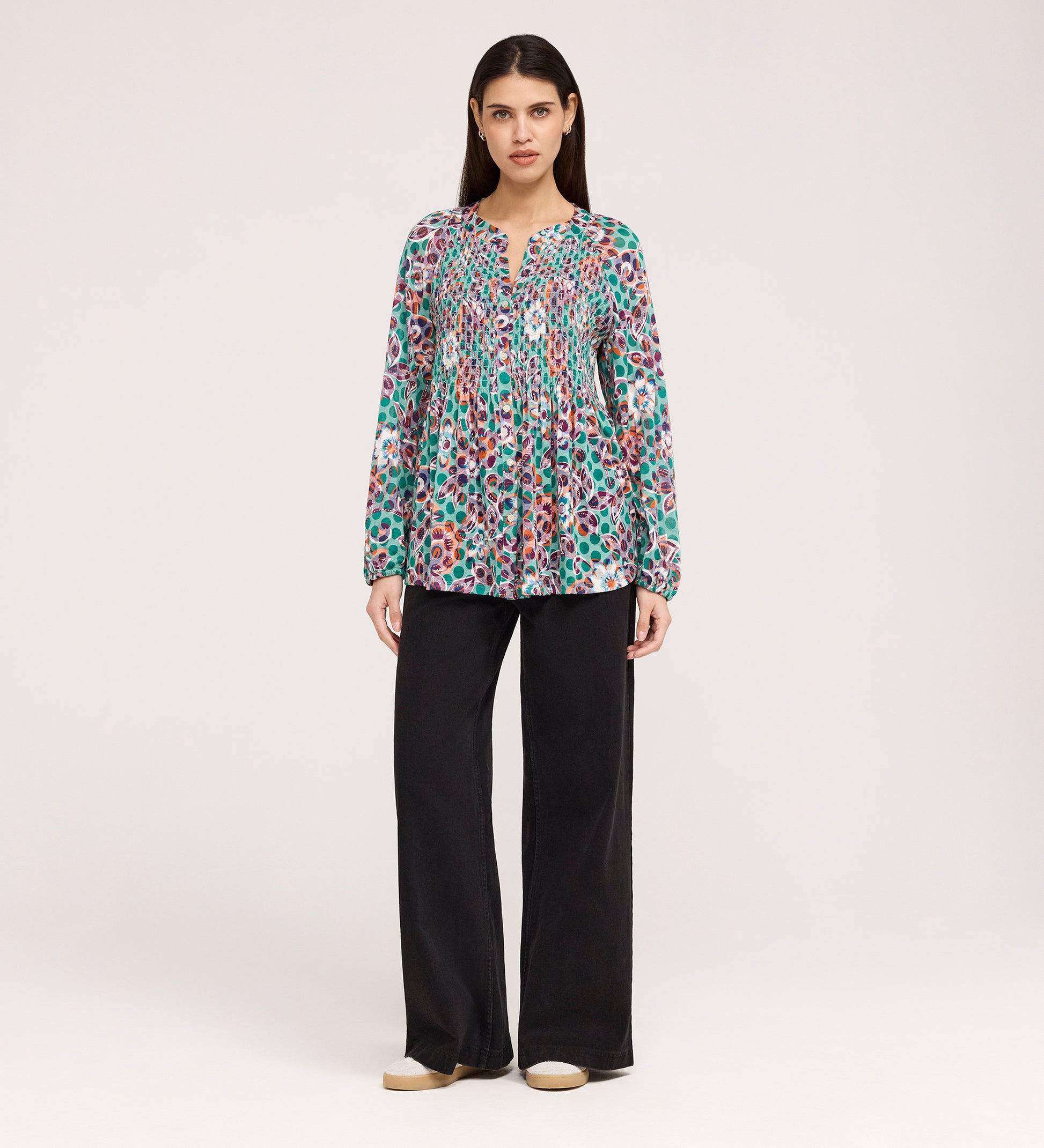 A woman stands facing forward in the Martha Green Floral Jersey Blouse with textured shirring, loose black flare-hem pants, and beige shoes, set against a plain light background.