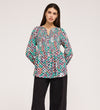 A woman stands with her hands in her pockets, wearing a Martha Green Floral Jersey Blouse with textured shirring and a split neckline, paired with loose black pants against a plain light background.