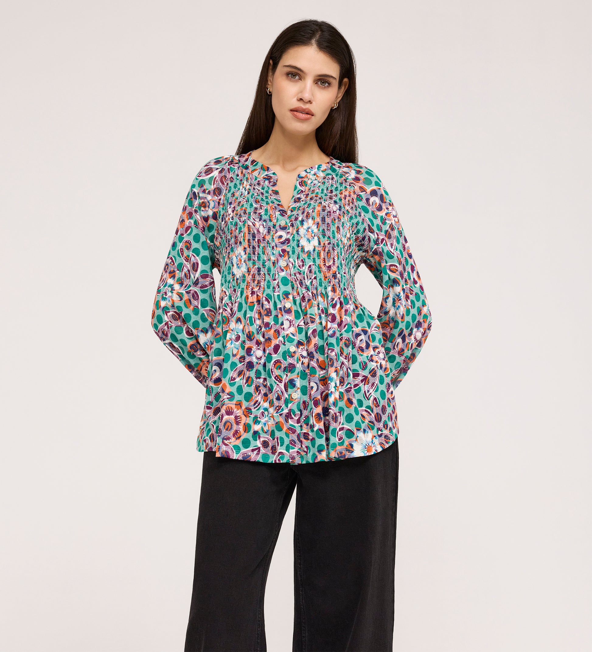 A woman stands with her hands in her pockets, wearing a Martha Green Floral Jersey Blouse with textured shirring and a split neckline, paired with loose black pants against a plain light background.