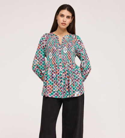 A woman stands with her hands in her pockets, wearing a Martha Green Floral Jersey Blouse with textured shirring and a split neckline, paired with loose black pants against a plain light background.