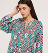 A woman with long brown hair wears the Martha Green Floral Jersey Blouse, featuring a flare hem and textured shirring. She looks down, one hand touching her head, against a plain light background.