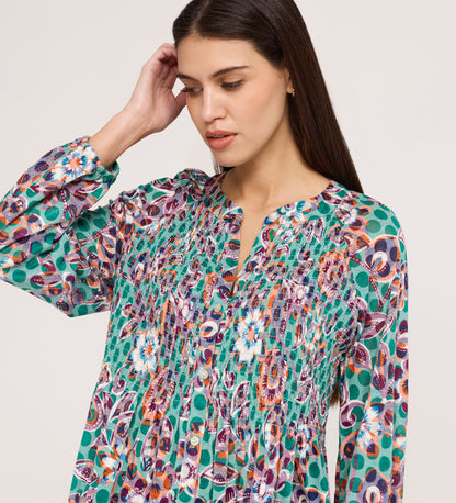 A woman with long brown hair wears the Martha Green Floral Jersey Blouse, featuring a flare hem and textured shirring. She looks down, one hand touching her head, against a plain light background.