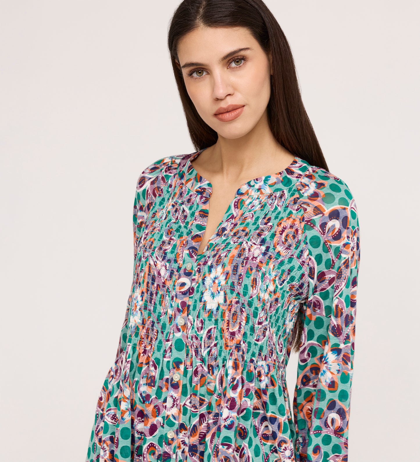 Woman with long dark hair wearing the Martha Green Floral Jersey Blouse, which features a colorful, patterned design with textured shirring and a split neckline, standing against a light background and looking slightly to the side.
