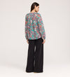 A woman stands facing away, wearing a vibrant Martha Green Floral Jersey Blouse with textured shirring and wide-leg black pants against a plain, light-colored background.