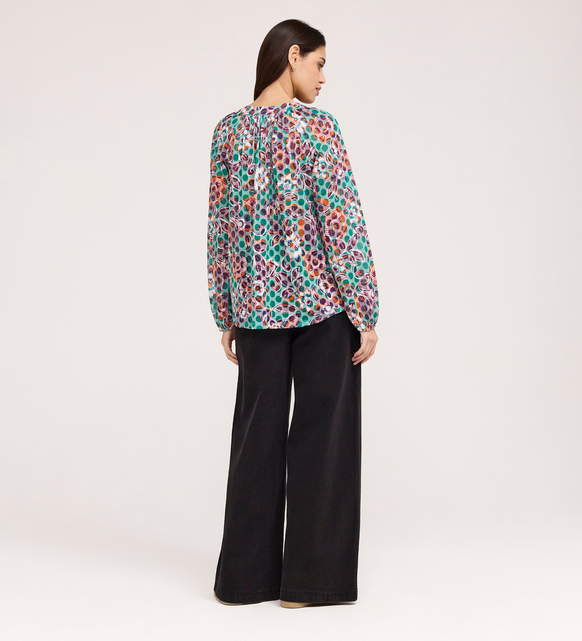 A woman stands facing away, wearing a vibrant Martha Green Floral Jersey Blouse with textured shirring and wide-leg black pants against a plain, light-colored background.