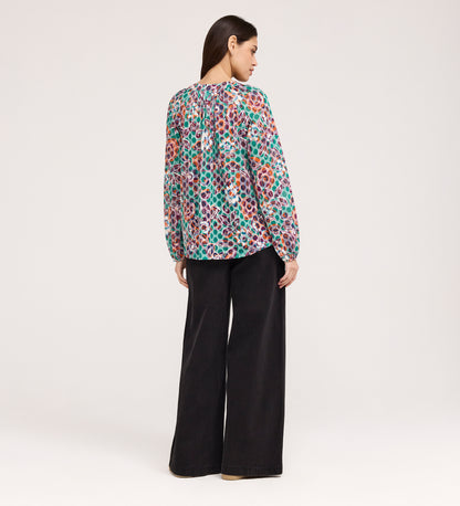 A woman stands facing away, wearing a vibrant Martha Green Floral Jersey Blouse with textured shirring and wide-leg black pants against a plain, light-colored background.