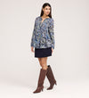 A woman poses against a light background, wearing the Martha Navy Floral Jersey Blouse with long sleeves and textured shirring, paired with a short navy skirt and knee-high brown boots. She stands slightly to the side, one hand by her hip.