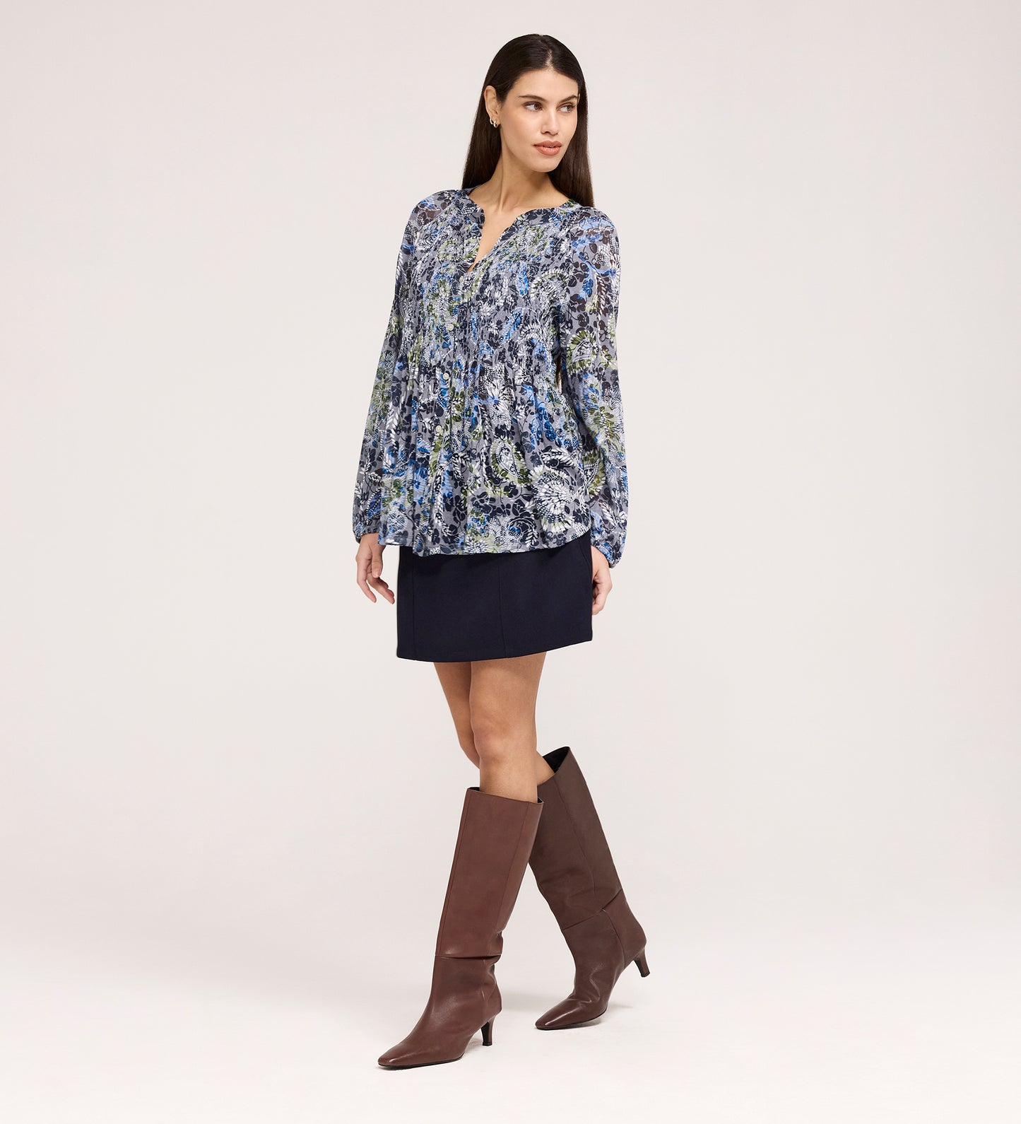 A woman poses against a light background, wearing the Martha Navy Floral Jersey Blouse with long sleeves and textured shirring, paired with a short navy skirt and knee-high brown boots. She stands slightly to the side, one hand by her hip.
