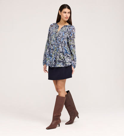 A woman poses against a light background, wearing the Martha Navy Floral Jersey Blouse with long sleeves and textured shirring, paired with a short navy skirt and knee-high brown boots. She stands slightly to the side, one hand by her hip.
