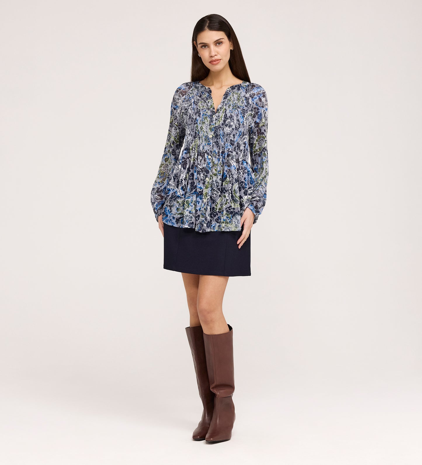 A woman stands against a plain background wearing the Martha Navy Floral Jersey Blouse, a dark mini skirt, and knee-high brown boots. She faces forward with her hands relaxed by her sides.