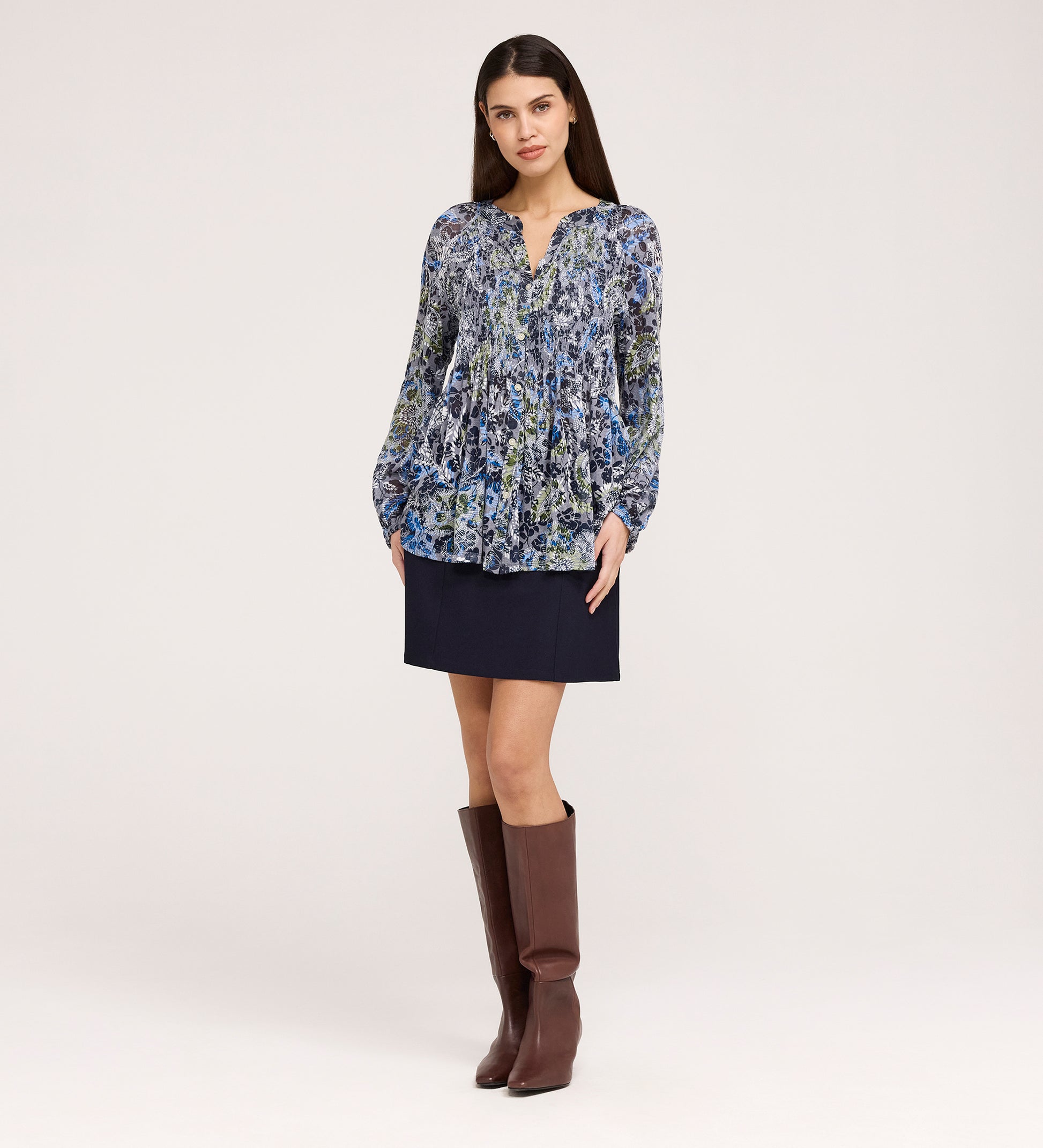 A woman stands against a plain background wearing the Martha Navy Floral Jersey Blouse, a dark mini skirt, and knee-high brown boots. She faces forward with her hands relaxed by her sides.