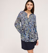 A woman with long brown hair wears the Martha Navy Floral Jersey Blouse, featuring blue, green, and black florals and shirring, paired with a navy skirt with a subtle flare hem. She stands against a plain light background, looking to the side.