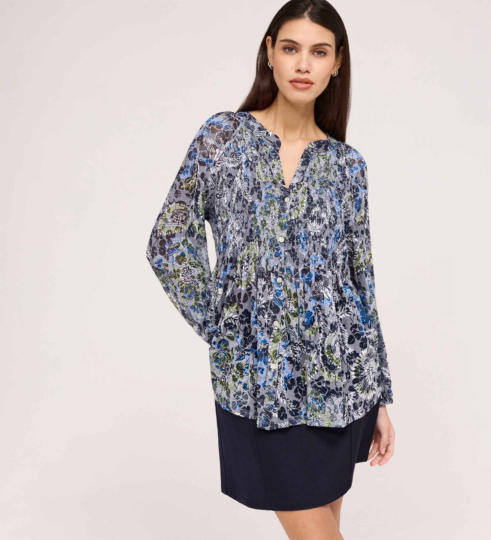 A woman with long brown hair wears the Martha Navy Floral Jersey Blouse, featuring blue, green, and black florals and shirring, paired with a navy skirt with a subtle flare hem. She stands against a plain light background, looking to the side.