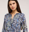 A woman with long dark hair wears the Martha Navy Floral Jersey Blouse, featuring blue, grey, green, and black floral patterns and textured shirring, as she looks at the camera against a neutral background.