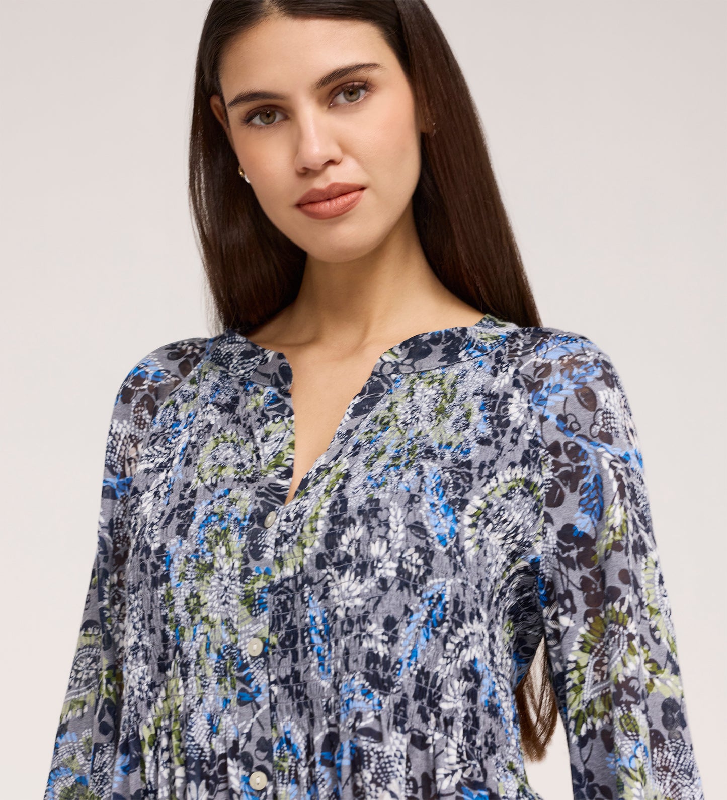 A woman with long dark hair wears the Martha Navy Floral Jersey Blouse, featuring blue, grey, green, and black floral patterns and textured shirring, as she looks at the camera against a neutral background.