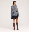 A woman stands facing away, wearing a Martha Navy Floral Jersey Blouse with long sleeves, a dark short skirt, and tall brown boots against a plain light background.