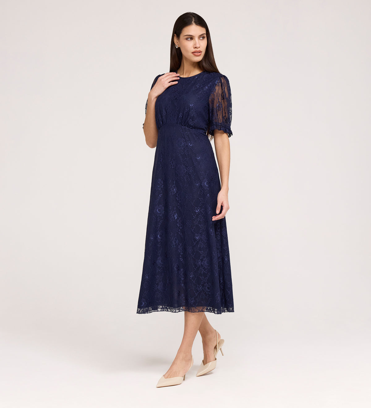 A woman poses in the Mela Navy Lace Midi Dress, featuring a knee-length lace design with a round neckline and short sleeves, paired with beige pointed heels against a plain light background.