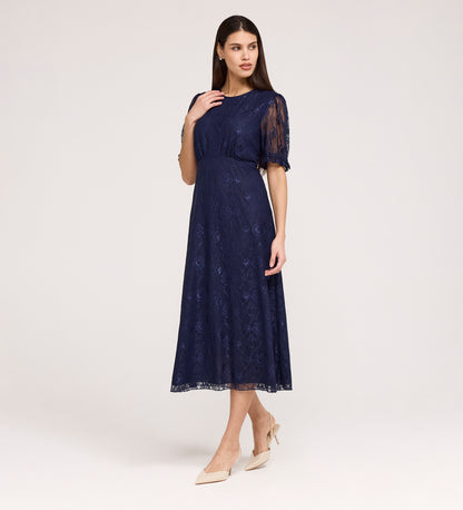 A woman poses in the Mela Navy Lace Midi Dress, featuring a knee-length lace design with a round neckline and short sleeves, paired with beige pointed heels against a plain light background.