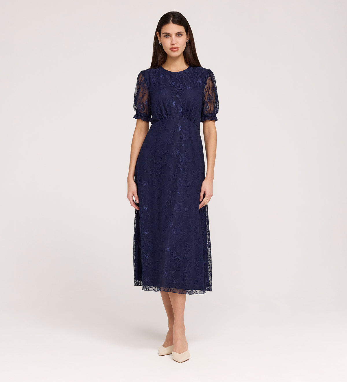 A woman with straight dark hair and a neutral expression models the Mela Navy Lace Midi Dress, featuring an elegant round neckline and short puffed sleeves, paired with beige pointed-toe shoes against a plain backdrop.
