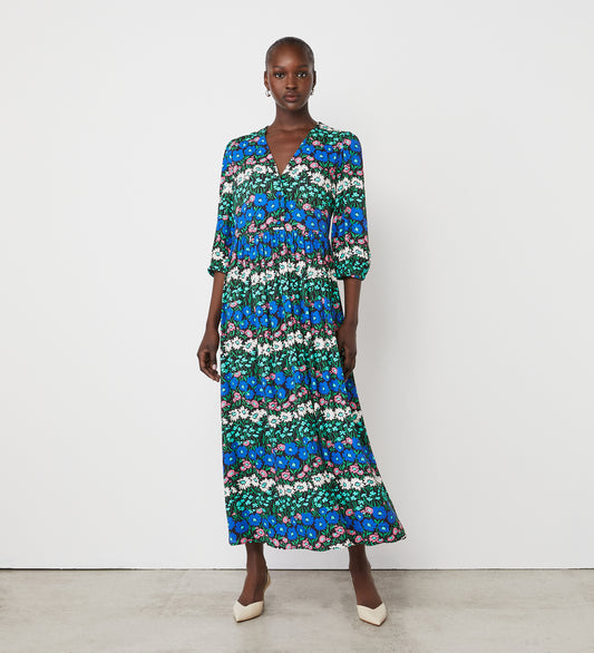 A woman stands against a white wall, wearing the Naomi Blue Floral Stripe Crepe Midi Dress—a long midi dress with three-quarter sleeves and a vibrant green, blue, pink, and white floral stripe pattern—paired with white pointed-toe heels.