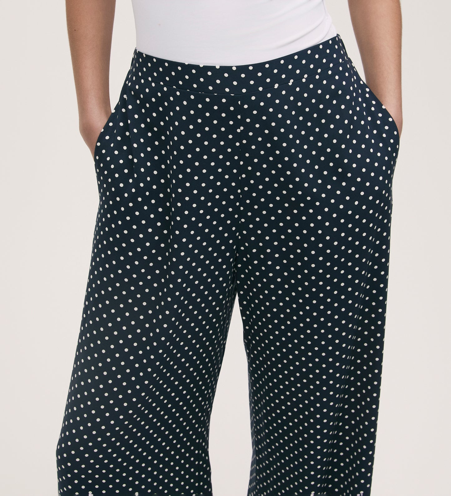Someone wears the Nat Navy Polka Dot Wide Leg Trousers with an elasticated waist, styled with a white fitted top and hands in the pockets.
