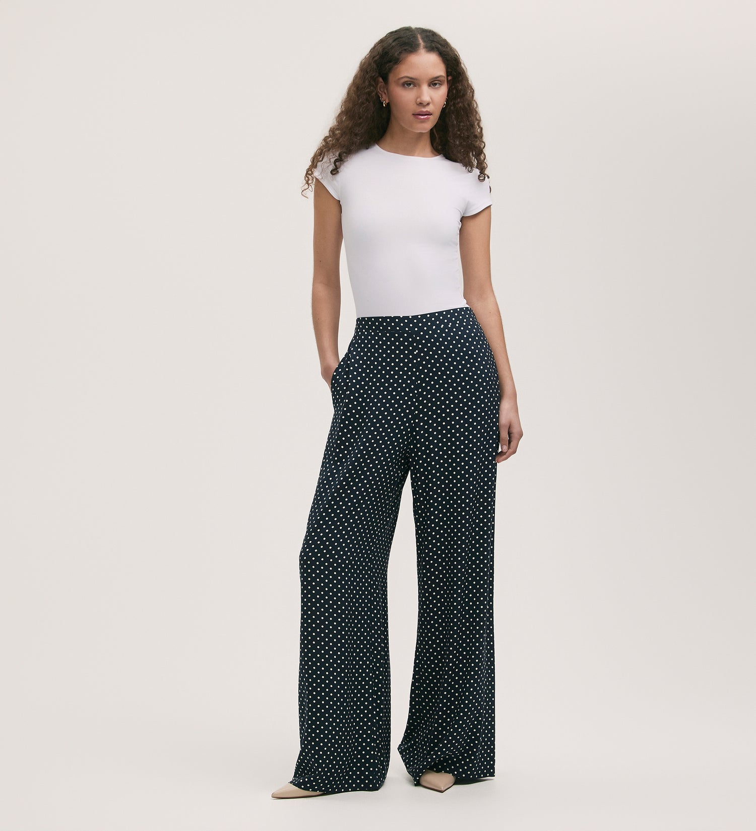 A woman with long curly hair wears a fitted white short-sleeve top and the Nat Navy Polka Dot Wide Leg Trousers, which are high-waisted with an elasticated waist. She stands against a plain light background with her hands in her pockets.