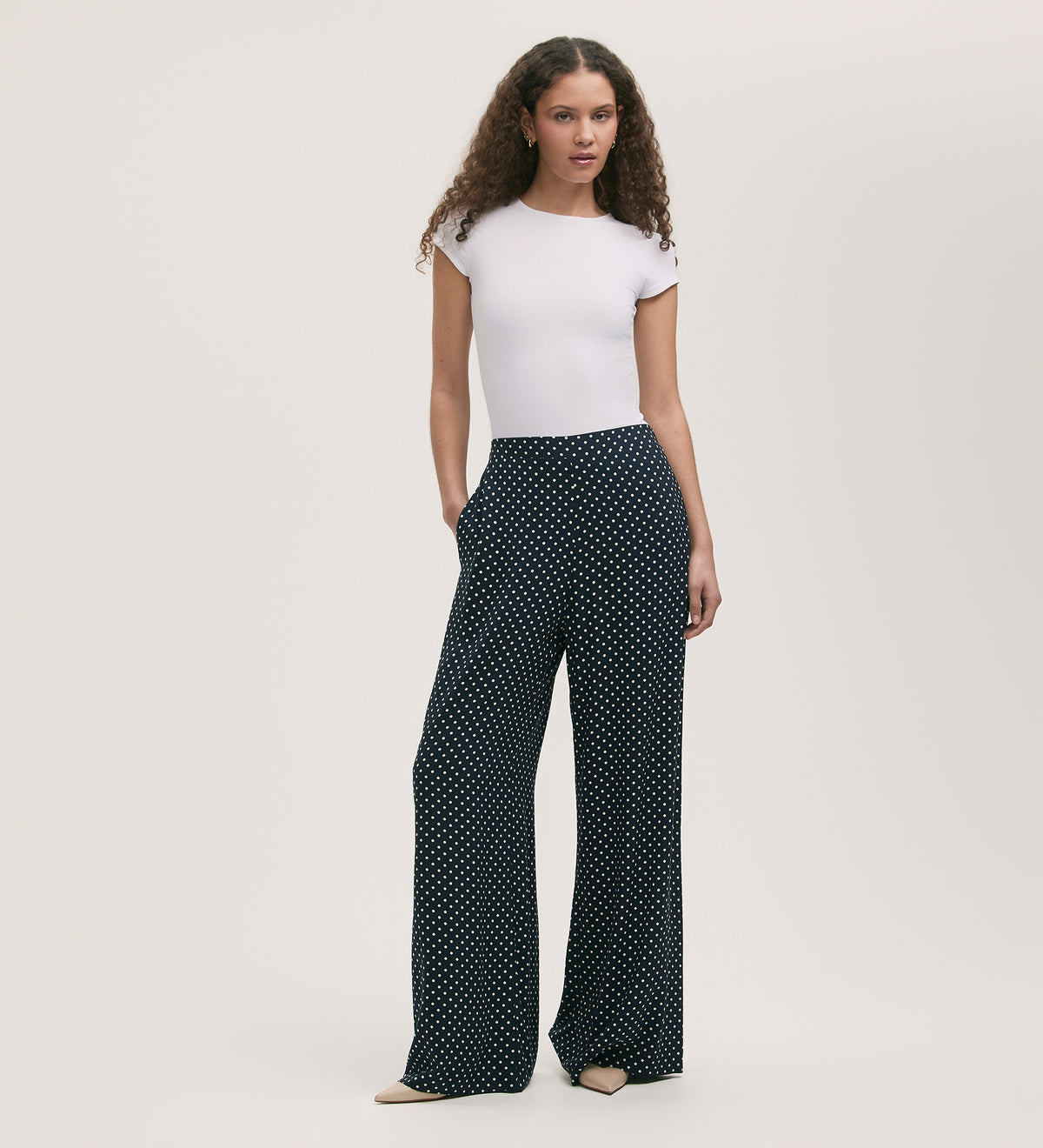 A woman with curly hair wears a fitted white short-sleeve top and the Nat Navy Polka Dot Wide Leg Trousers with an elasticated waist, paired with beige pointed-toe shoes, standing against a plain light background.