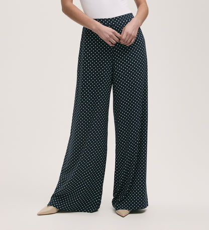 A person wearing Nat Navy Polka Dot Wide Leg Trousers with an elasticated waist and beige pointed-toe shoes, standing with hands clasped in front.