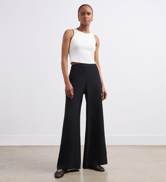 A woman stands against a white wall, wearing a sleeveless white crop top and the Nat Black Crepe Wide Leg Trousers with functional pockets. She faces forward with minimal accessories and black flat shoes.