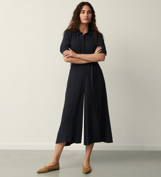 A woman with long, wavy hair stands against a plain wall, confidently wearing the Floralie Black Jumpsuit— a loose-fitting, short-sleeve black satin jumpsuit with wide leg trousers and a waist tie. Her arms are crossed as she looks at the camera.