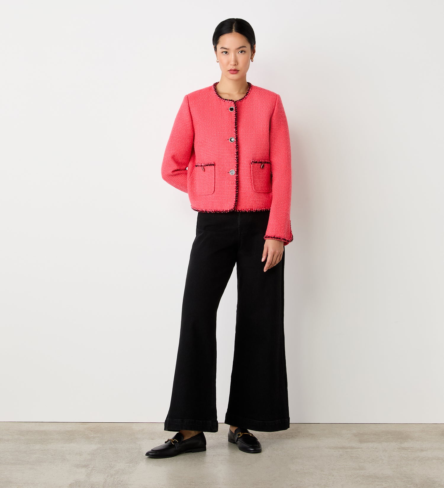 A woman stands against a plain white wall wearing the Polly Coral Jacket, a bright pink cropped style with contrast trim and front pockets, paired with black wide-leg pants and black loafers. Her hair is tied back and she has a neutral expression.