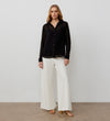 A woman stands against a plain background wearing the Janette Black Chiffon Blouse With Detachable Cami— a sheer black button-up with textured spot detail—paired with white wide-leg pants and gold heeled sandals.